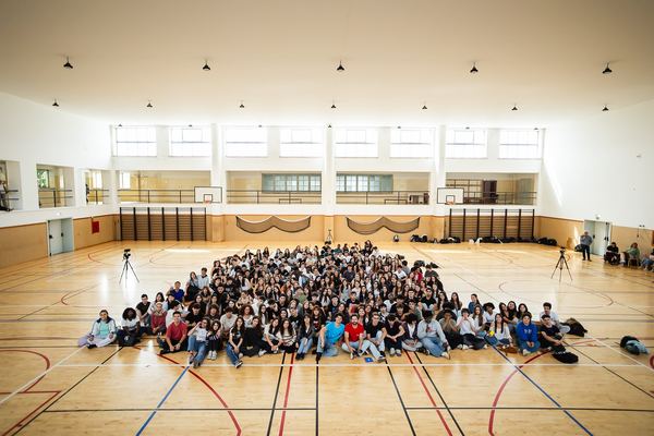 Plano largo da assistência: centenas de alunos sentados no pavilhão.