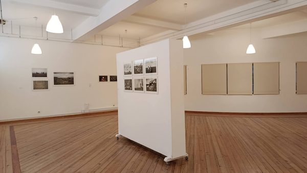 Vista da Sala de Exposições vazia: um painel branco ao centro com seis fotografias a preto e branco e, ao fundo, outras fotografias emolduradas na parede.