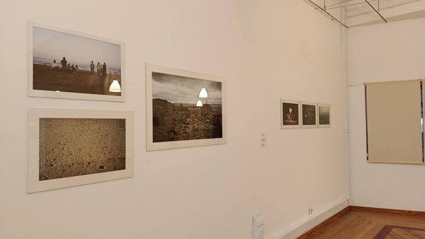 Pormenor de uma parede da exposição com três fotografias a cores de grande formato — uma praia com pessoas ao longe, um areal e uma paisagem rochosa — e, ao lado, três fotografias mais pequenas.
