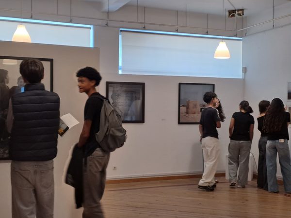 Alunos a visitar a exposição, de pé, a observar as fotografias emolduradas nas paredes da sala.