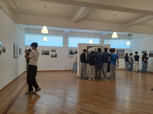 Um grupo de alunos reunido em frente a um painel com fotografias, ao centro da Sala de Exposições, e outros a percorrer a sala.