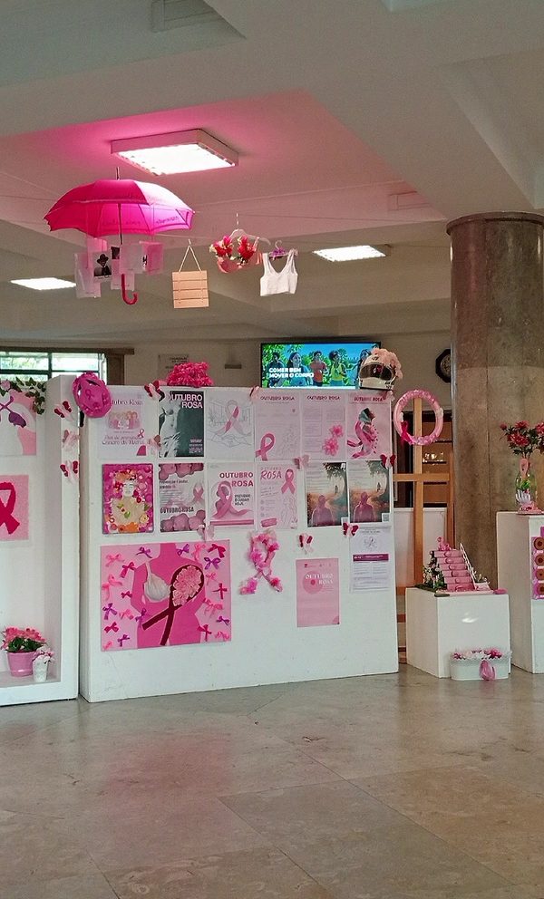 Vista do átrio da escola decorado para o Outubro Rosa: painéis brancos cobertos de cartazes e trabalhos cor-de-rosa, um guarda-chuva rosa e peças de roupa penduradas do teto e plintos com flores.