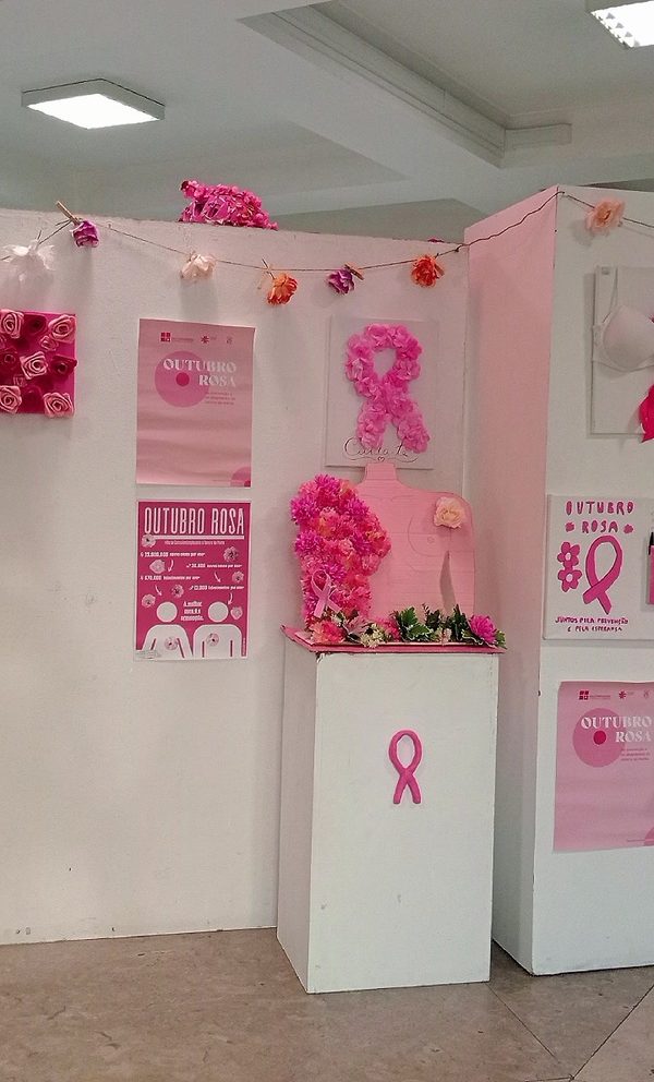 Painéis da exposição com cartazes «Outubro Rosa», um laço feito de flores de papel, uma grinalda de flores e, num plinto branco com um laço rosa, um busto coberto de flores cor-de-rosa.