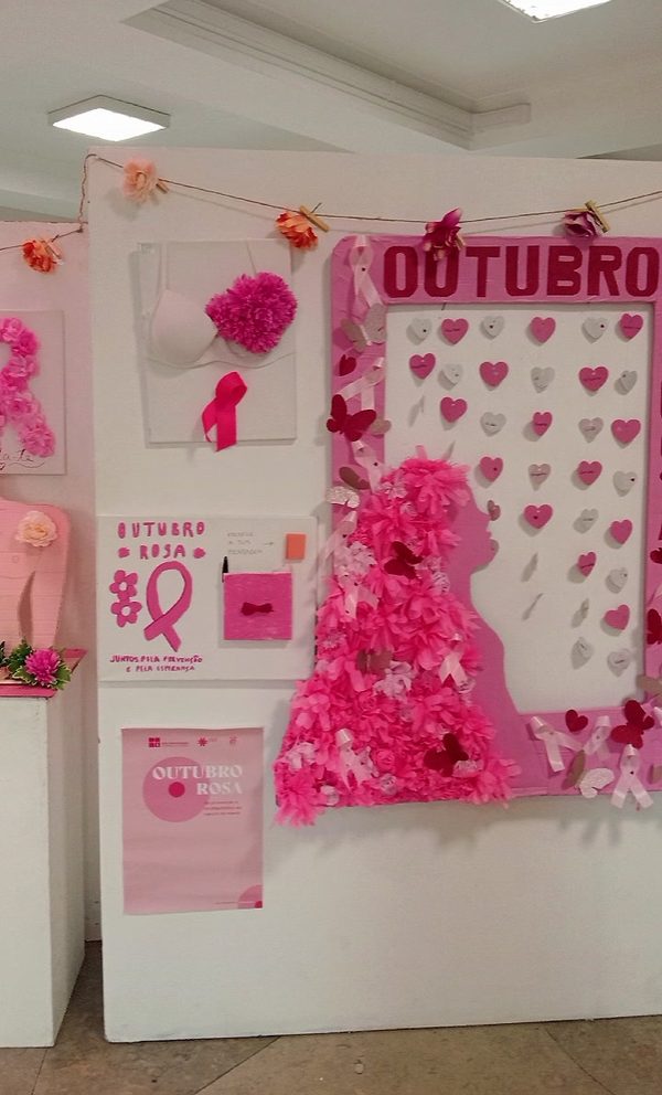 Painel com a palavra «OUTUBRO» em letras cor-de-rosa, um quadro de corações e a silhueta de um vestido feita de flores cor-de-rosa; ao lado, cartazes com o laço rosa.