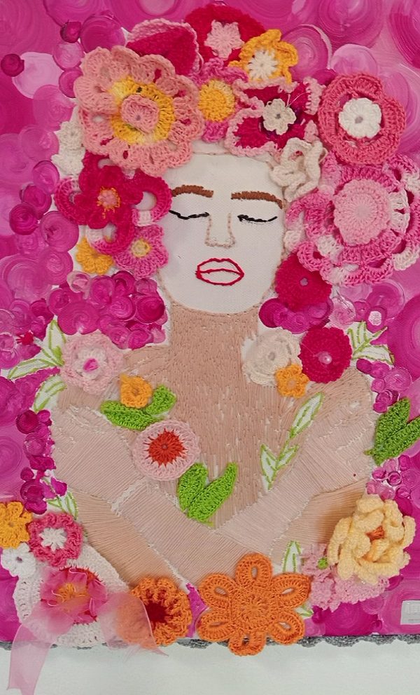 Trabalho em croché: o rosto de uma mulher de olhos fechados, com o cabelo feito de dezenas de flores de croché em rosa, laranja e amarelo.