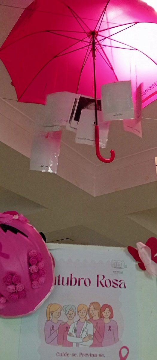 Guarda-chuva cor-de-rosa aberto, suspenso do teto do átrio, com mensagens em papel penduradas; por baixo, um cartaz «Outubro Rosa» com ilustrações de mulheres e a frase «Cuide-se. Previna-se.»