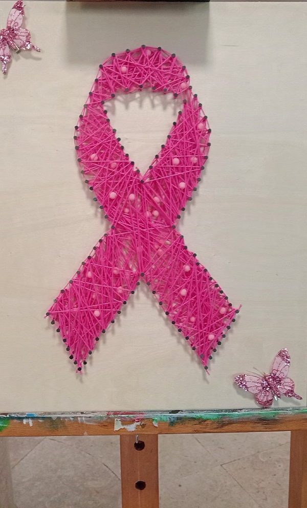 Laço cor-de-rosa feito com fio esticado entre pregos sobre uma placa clara, num cavalete, com duas borboletas decorativas.